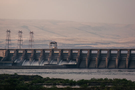 View of The Dalles Dam on the Columbia River with concrete spillways, power transmission towers, and rolling hills in the background in The Dalles, Oregon, United States.