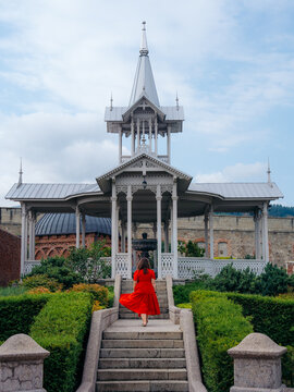 View of a woman in a red dress walking up stone stairs towards an ornate white wooden pavilion at Rabati Fortress in Akhaltsikhe, Samtskhe-Javakheti, Georgia.