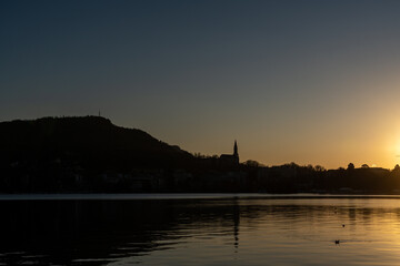 Naklejka premium Silhouette of a town with a church spire and hills at sunset, viewed across a calm lake with reflections.