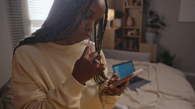 African American young woman brushing teeth while scrolling on smartphone in bedroom. Footage suitable for commercial use in digital burnout and screen addiction campaigns