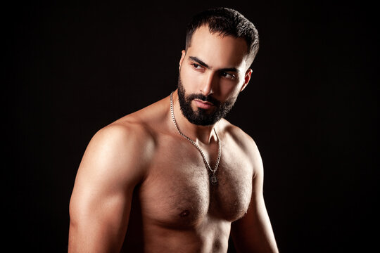 Shirtless fit man with well-defined muscular shoulders and pectorals in relaxed pose wearing silver chain necklace in dramatic lighting isolated over black background