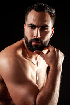 Muscular man touching chin posing with strong arm showing defined biceps and triceps muscles wearing silver chain jewelry in studio lighting isolated over black background