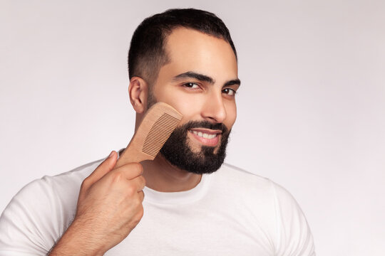 Man brushing thick black beard with wooden tool to create perfect shape and smooth texture for masculine grooming procedure isolated over gray background