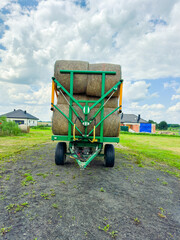 Farm equipment carries large hay bales under a cloudy sky near rural buildings during the day © DSMT