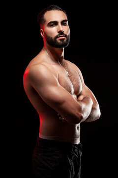 Strong guy standing sideways showing massive bicep and triceps with crossed arms and athletic torso highlights by red glow isolated over black background