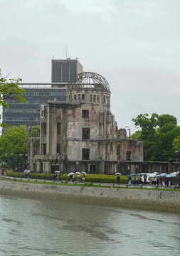 View of the historic Genbaku Dome Atomic Bomb Dome with its iconic metal frame and brick ruins, situated along the Motoyasu River with people walking under umbrellas in Hiroshima, Hiroshima, Japan.