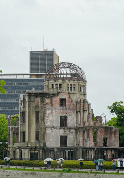 View of the historic Atomic Bomb Dome with its iconic metal frame and weathered brick walls under an overcast sky, with people walking by holding umbrellas in Hiroshima, Hiroshima, Japan.