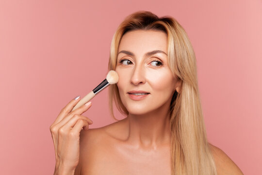Mature woman looking aside while applying makeup brush focusing on flawless skin and delicate facial contours beauty concept isolated on pink background