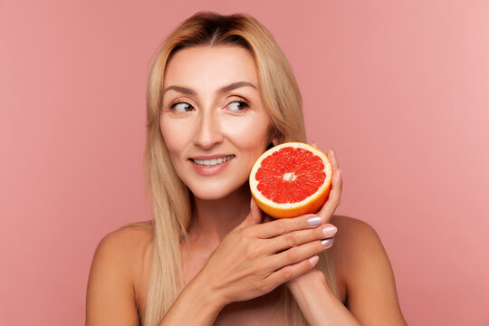 Cheerful blonde lady holding fresh grapefruit slice for organic skincare and healthy vitamin nutrition isolated on pink background