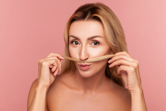 Playful woman making a mustache with her blonde hair lock while posing for creative beauty campaign isolated on pink background