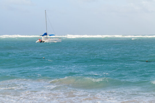 An anchored catamaran sailboat drifts on turquoise ocean waters near a sandy beach, framed by gentle waves and a distant horizon, evoking relaxed tropical sailing, travel and seaside leisure
