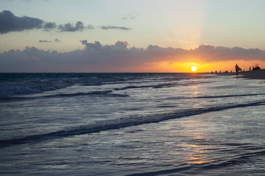 Golden sunset glows on the horizon as gentle waves wash a reflective shoreline. Silhouetted figures stroll the sandy beach, creating a peaceful, romantic and contemplative coastal scene at dusk