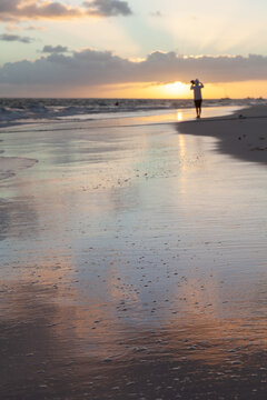 A solitary figure walks along a tranquil beach at sunset, silhouetted by golden light while colorful clouds and wet sand reflect the warm sky, suggesting calm, solitude and peaceful contemplation