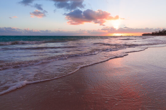 Golden-pink sunset over a tropical beach with gentle waves washing the wet sand, soft clouds and distant palms creating a calm, romantic and meditative coastal atmosphere for travel and relaxation