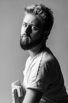 Monochrome bearded man looking directly at camera with intense and serious expression sitting in white t-shirt leaning on wooden chair isolated on gray background
