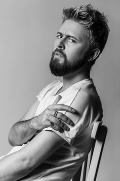 Monochrome intense man looking at camera over his shoulder leaning on wooden chair with confident and strong gaze isolated on gray background