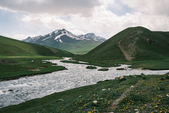 View of a winding river flowing through a lush green valley with snow-capped mountains under a cloudy sky in Kyrgyzstan.