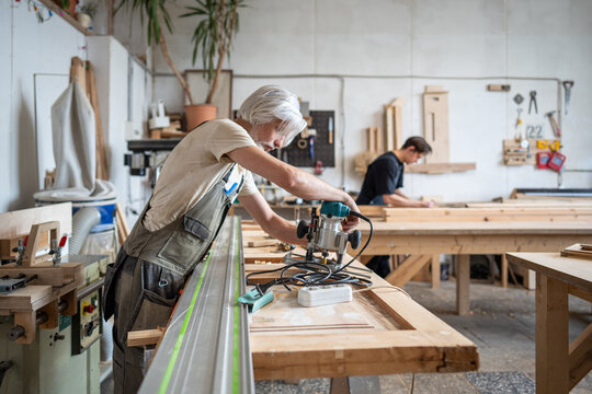 Woodworker engineer preparing electric wood router machine at workstation in restoration workshop, skilled craftsman technician adjusting instrument for antique wooden door reconstruction in studio.