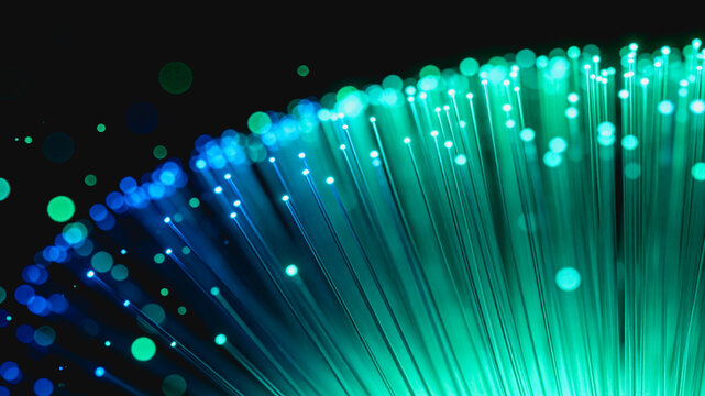 Glowing blue and green fiber optic strands emit light particles against a dark background, representing digital data transmission.