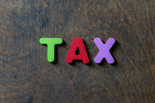 Word tax written with colorful toy letters on dark wooden background. Top view closeup of word tax spelled out in green, red and purple wooden blocks centered on rustic scratched wood table surface
