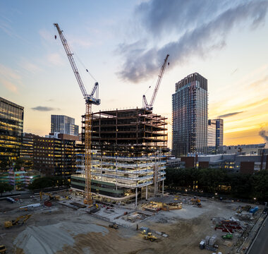 Aerial view of a construction site with steel framework, towering cranes, and surrounding urban buildings during a golden hour sunset in Cambridge, Massachusetts, United States.