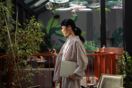 Woman walking in cafe looking at phone holding laptop