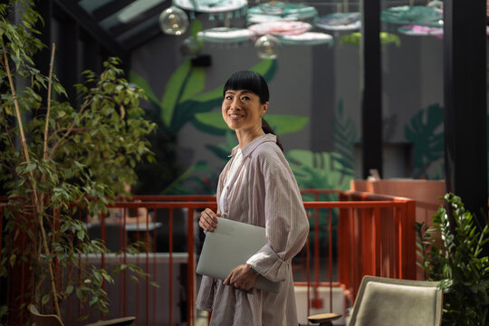 Asian businesswoman smiling holding laptop in modern co-working space