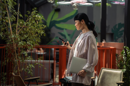 Young woman using smartphone holding laptop in modern workspace