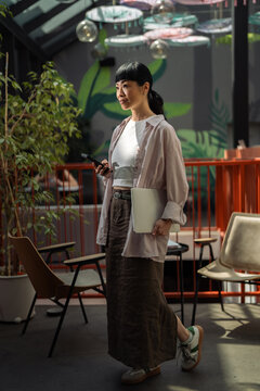 Asian woman walking in a cafe holding smartphone and laptop
