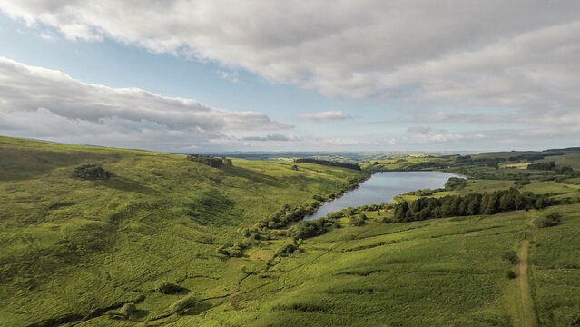 Aerial view of Cray Reservoir surrounded by rolling green hills and lush meadows under a cloudy sky in Brecon, Wales, United Kingdom.