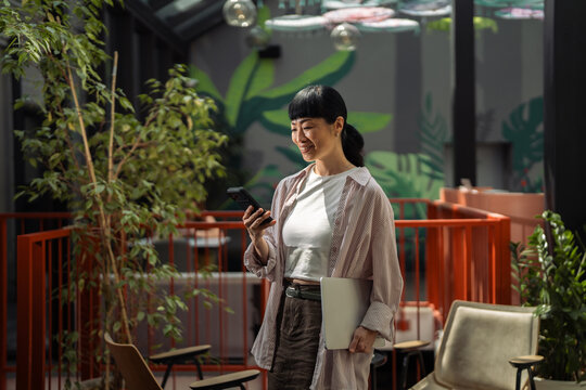 Asian woman smiling using smartphone holding laptop