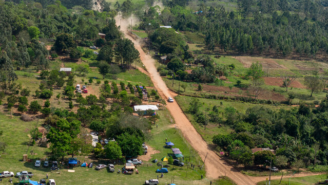 Aerial view of a rally car driving on a dusty dirt road through a rural landscape with trees, fields, and parked spectator vehicles in Nueva Alborada, Itapua Department, Paraguay.