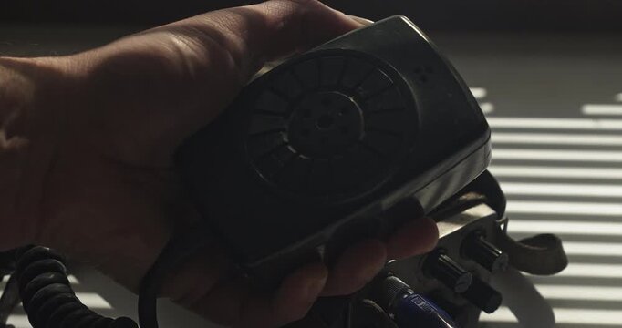 Close up of a police detective keying the microphone button on a vintage walkie talkie in a low-lit room, suggesting surveillance and covert communication.
