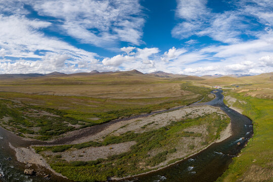 Aerial view of a winding river flowing through the vast, grassy plains and rolling hills under a blue sky with scattered clouds in Gilgit-Baltistan, Pakistan.
