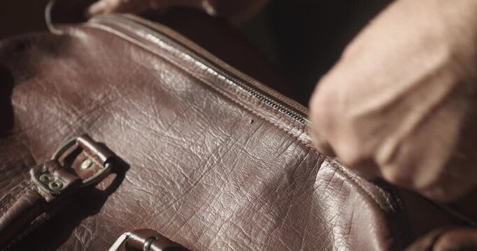 Close-up of hands zipping a well used leather travel bag closed in low cinematic lighting, evoking nostalgic travel and departure.