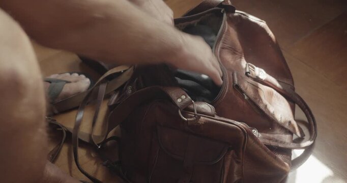 Person wearing shorts and flip flops packs personal items and binoculars into a leather holdall while preparing for a nostalgic travel adventure.