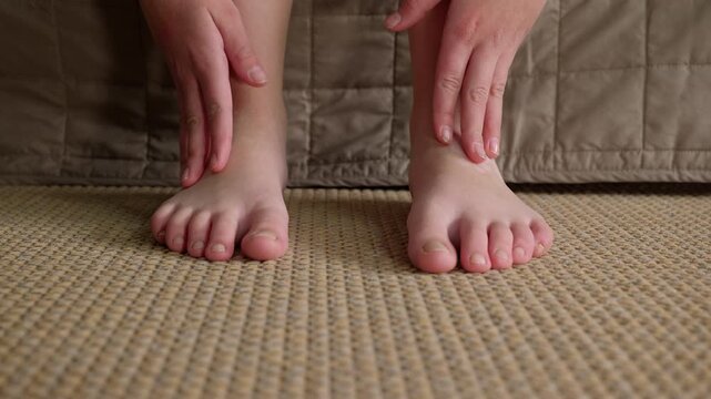 Person massaging and scratching their itchy feet. Unrecognizable person sitting on a bed and scratching their ankles and massaging their tired feet due to an allergic reaction, irritation