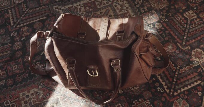 Old leather travel bag is dropped onto a patterned carpet as sunlight highlights the scene, evoking nostalgic travel and returning home.