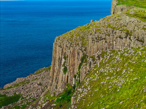 Aerial view of Fair Head cliffs with a climber on the rock face, green grassy slopes, and rocky terrain overlooking the deep blue sea in Ballycastle, Northern Ireland, United Kingdom.