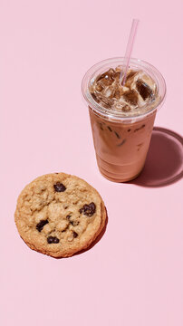 Iced coffee with milk in a takeaway cup beside a chocolate chip cookie on pastel pink background, refreshing caf? drink and sweet snack with copy space