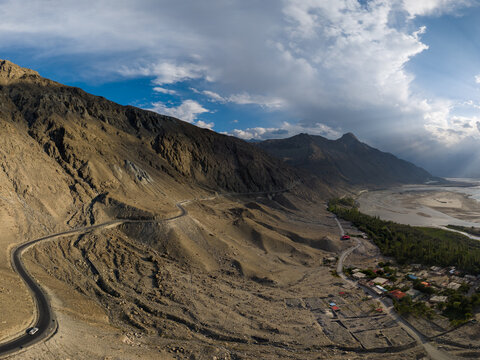 Aerial view of a winding mountain road traversing the rugged landscape near a river valley and a small settlement, Gilgit-Baltistan, Pakistan.