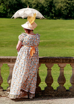 A woman dressed in a historical Regency-style floral gown and hat stands by a stone balustrade, shielding herself with an elegant lace parasol while enjoying a peaceful day in a garden setting.