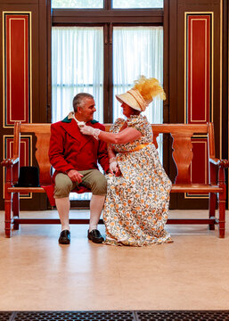 A man and a woman dressed in 19th-century historical attire sit on a wooden bench, where the woman affectionately adjusts the man's cravat during a historical reenactment or themed event.