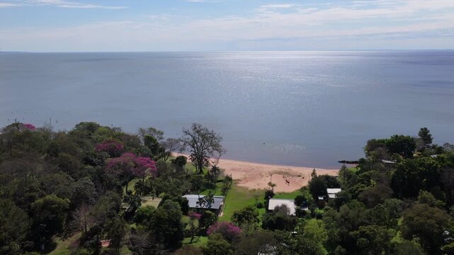 Parana River coastline and wooded Santa Tecla landscape in Corrientes, Argentina, with a sandy shore, houses and dense vegetation, drone forward flyover.