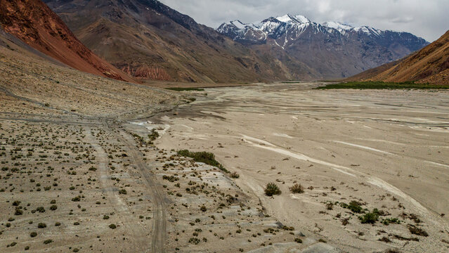 Aerial view of the rugged Bartang River valley featuring a wide, dry braided riverbed, sparse desert vegetation, and distant snow-capped mountains in Chah Ab, Gorno-Badakhshan Auto, Tajikistan.