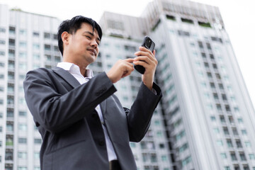 Fototapeta premium Focused Asian businessman using smartphone outdoors with modern office building blurred background. Professional corporate manager checking data on mobile phone during urban work commute in city