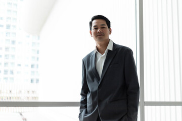 Fototapeta premium Confident Asian businessman in suit standing near office window looking at camera. Professional corporate executive posing with copy space inside modern high rise skyscraper building