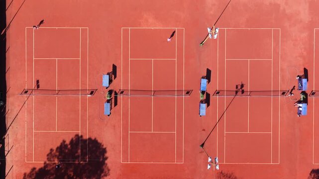 Top-down aerial view of multiple clay tennis courts with players creating geometric patterns during recreational matches.