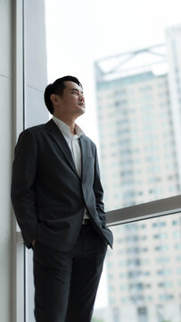 Confident Asian businessman in suit standing near glass window looking out at cityscape. Professional corporate executive thinking about future success in modern high rise office skyscraper
