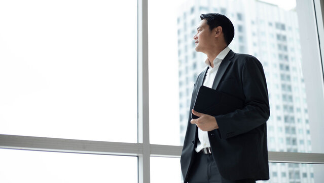 Visionary Asian businessman holding digital tablet while looking out window in modern office building. Thoughtful corporate executive planning future business strategy with copy space in skyscraper